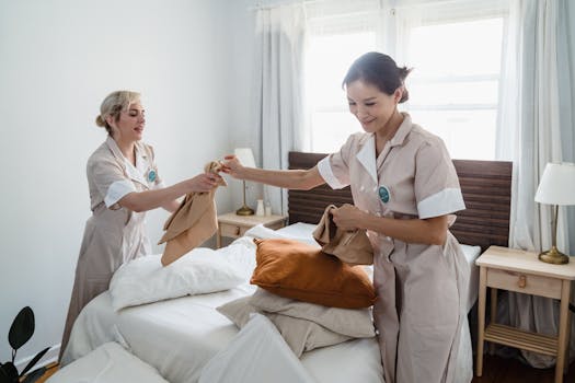 Two female housekeepers in uniform efficiently prepare a bed in a bright, well-lit room.