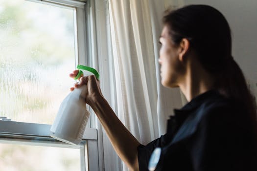 A woman using a spray bottle to clean a glass window inside a room, captured in a close-up shot.