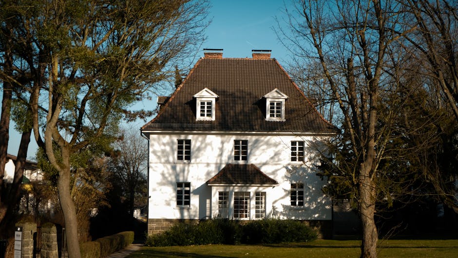 fenster putzer münchen - Elegant colonial-style house with traditional roof amid lush trees on a sunny day.