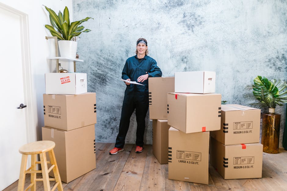 move out house cleaners - Smiling mover in coveralls standing with parcels, clipboard in hand, indoors.