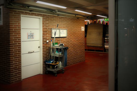 A brightly lit industrial hallway with a cleaning cart and supplies near an office door.